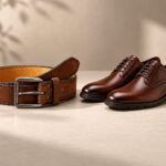 Brown leather belt with a silver buckle and matching formal shoes on a beige surface.