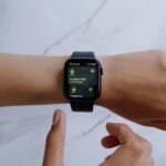 A hand wearing a black smartwatch displaying workout options: "Outdoor Walk" and "Indoor Run." Another hand is poised to select an option, suggesting focus.